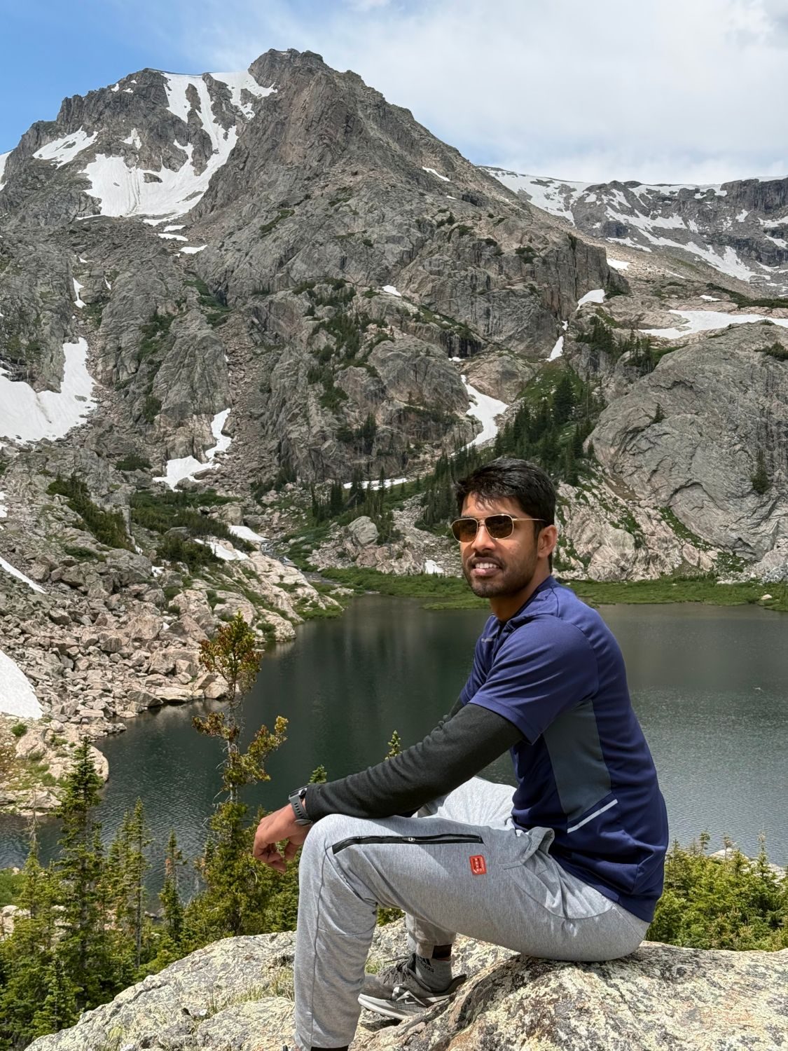 Abhishek sitting on a rock above an alpine lake below a snowy peak